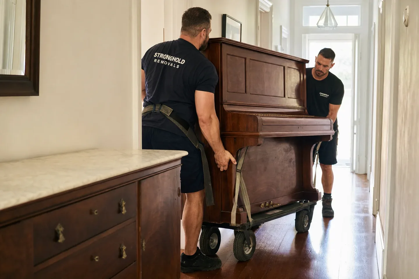 Removalists moving an upright piano on a trolley down a hallway